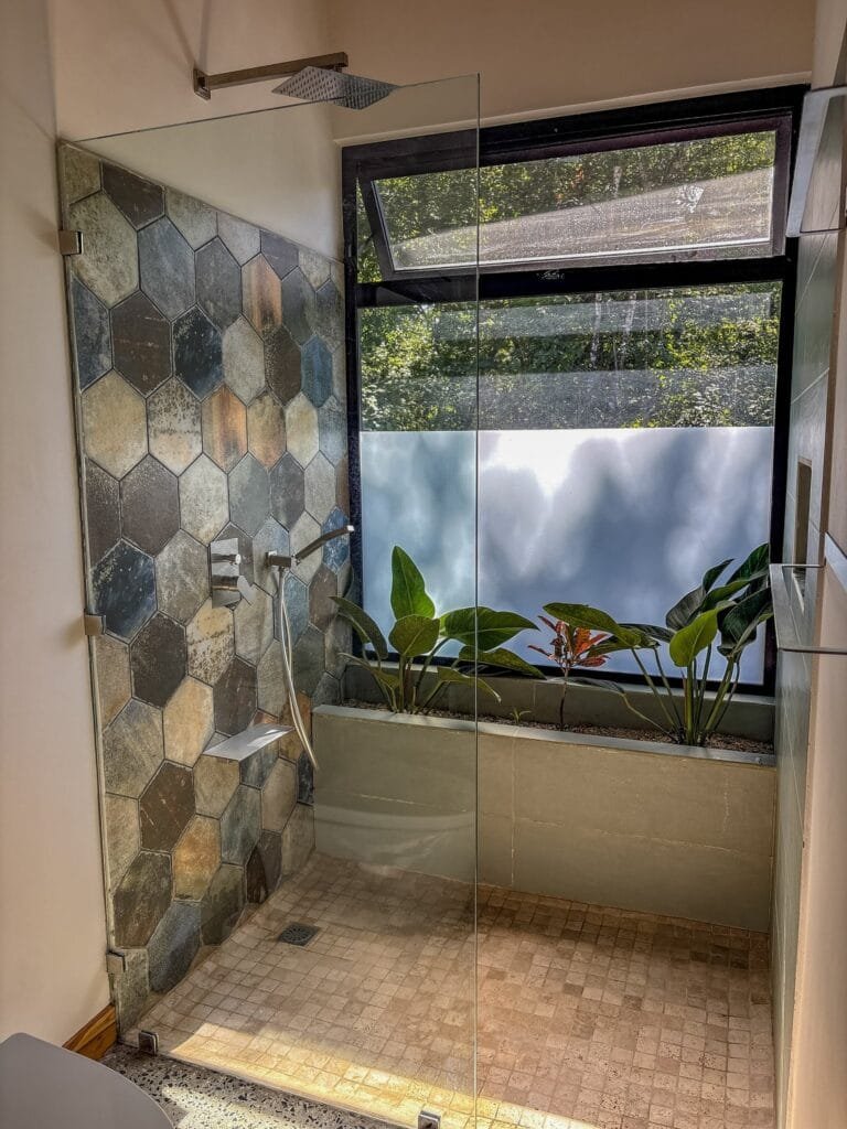 Ducha a ras de piso del apartamento Colibrí con azulejo hexagonal y plantas tropicales