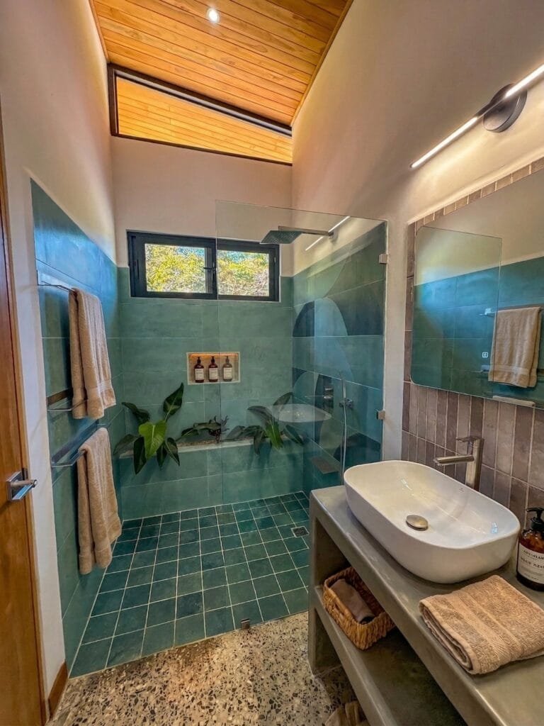 Ducha a ras de piso del apartamento Tucán con azulejo verde azulado y plantas tropicales